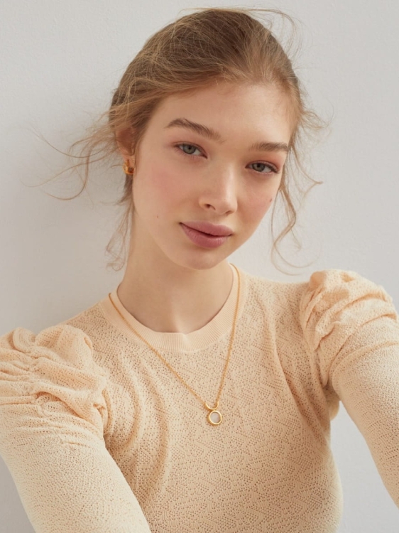 Modelka w beżowym swetrze prezentująca złoty naszyjnik z okrągłą zawieszką z kryształem górskim.