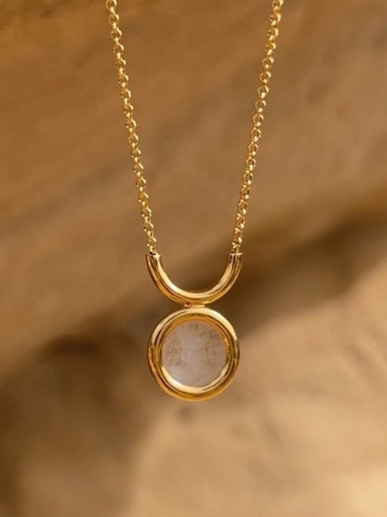 Złoty łańcuszek z kryształem górskim sfotografowany na piaskowym tle w ciepłym świetle.