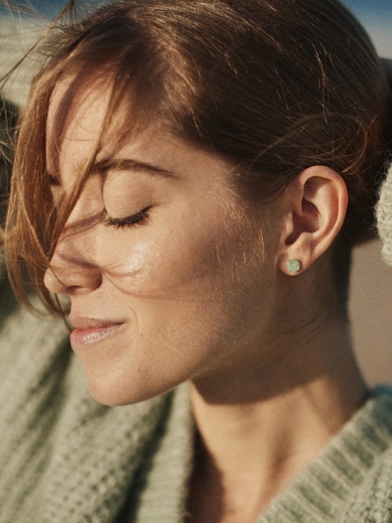 Zbliżenie na twarz modelki w kolczykach z zielonym kamieniem, uchwycona w ciepłym świetle.